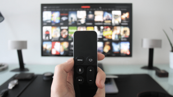 A hand holding a tv remote with tv on the background.
