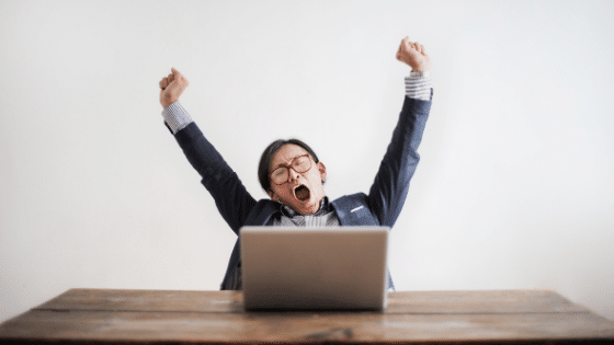 A man sitting down yawning and stretching in front of a laptop.