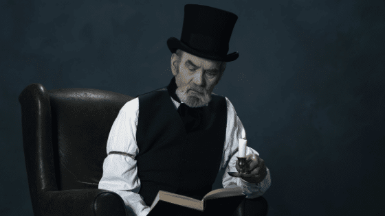A old man wearing a black hat sitting down reading a book while holding a lit candle.