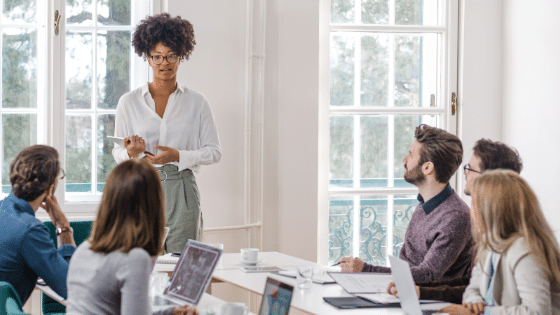 Business meeting with a young African woman speaking and standing while the rest listen